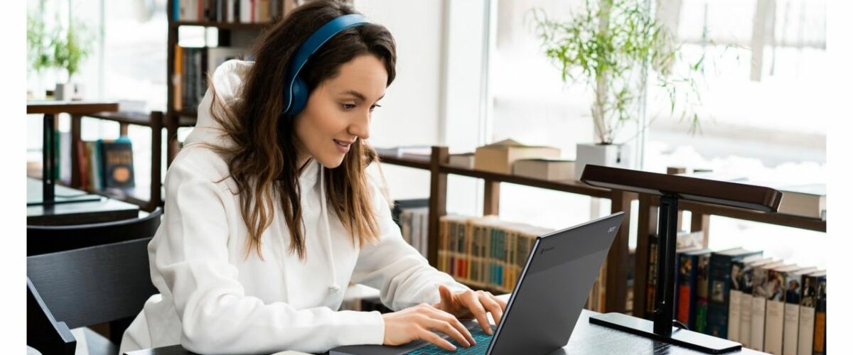 A student wearing headphones sitting at a wooden table with a laptop open in a library setting