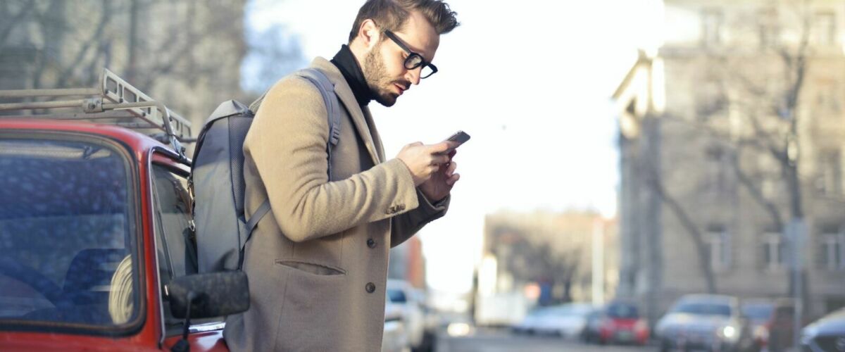 Man in Beige Coat Holding Phone Leaning on Red Vehicle