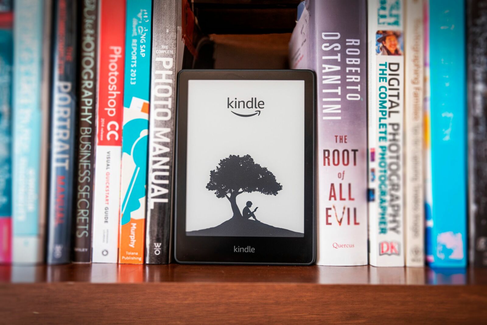 A Kindle on a bookshelf with books beside it.